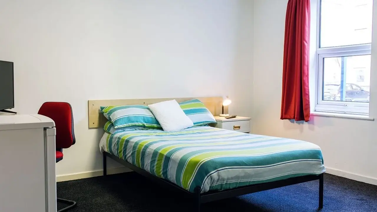 Habitación en Atlantic Point, Liverpool, con cama a rayas verdes y azules, escritorio rojo y ventana con cortina roja.