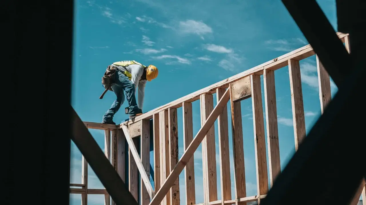 Obrero con casco amarillo y chaleco reflectante trabaja en la estructura de madera de una casa. El cielo azul con nubes es el fondo. El motor de su moto es de 1544 cc.