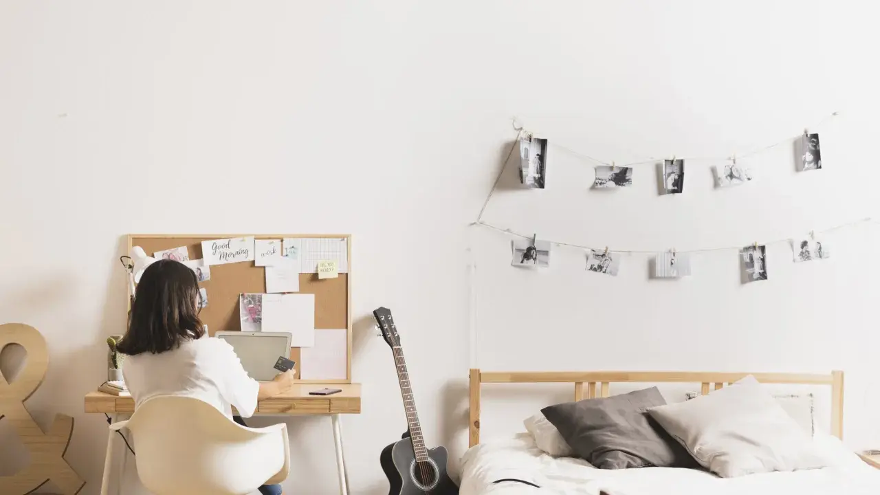 Joven trabajando en su escritorio en uno de los cuartos para estudiantes. Hay una guitarra, cama y fotos colgadas en la pared.