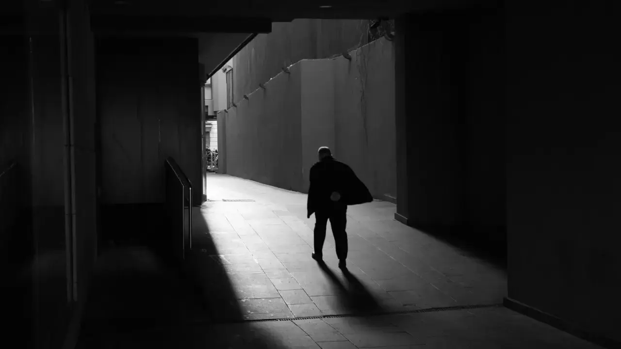 Hombre solitario camina por un pasillo oscuro, su figura se desvanece en la luz, un reflejo del precario significado de la existencia.