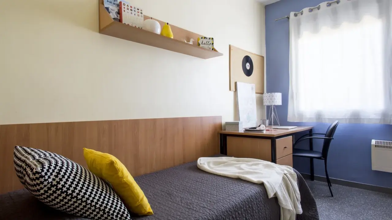 Habitación de estudiante en la carlos v residencia granada. Cama con cojines, escritorio con lámpara y estantería con libros.