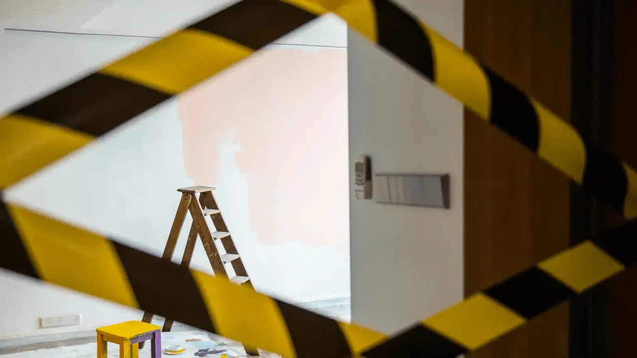 Escalera de madera y un taburete amarillo, listos para aplicar el **articulo 30 lau** en la pared. Cinta de precaución amarilla y negra.