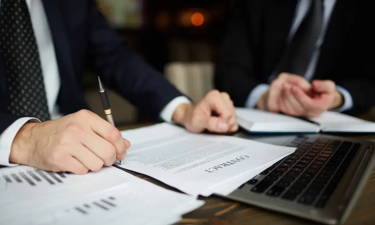Dos hombres de negocios revisan un contrato. Uno firma el documento, mientras el otro observa. Podría tratarse de un acuerdo legal, o quizás de un proceso de embargo por impago de alquiler.