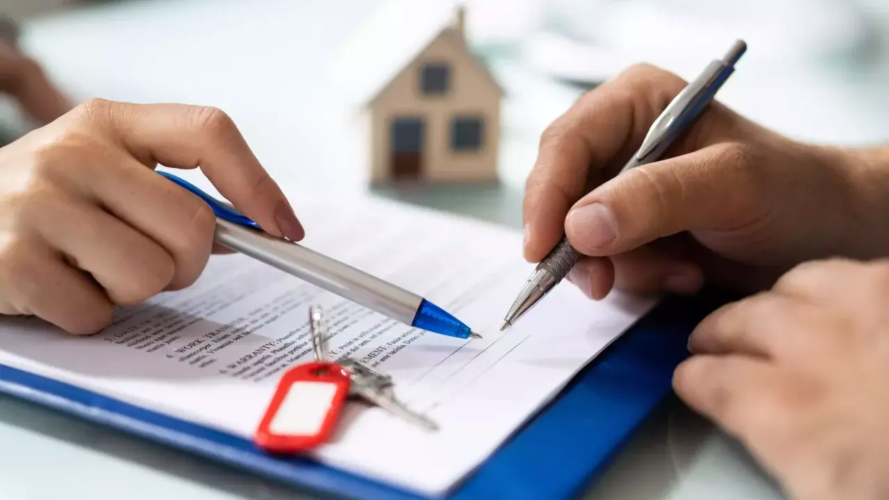 Manos firmando un documento, con llaves y una maqueta de casa. Podría ser un desistimiento de contrato de alquiler.