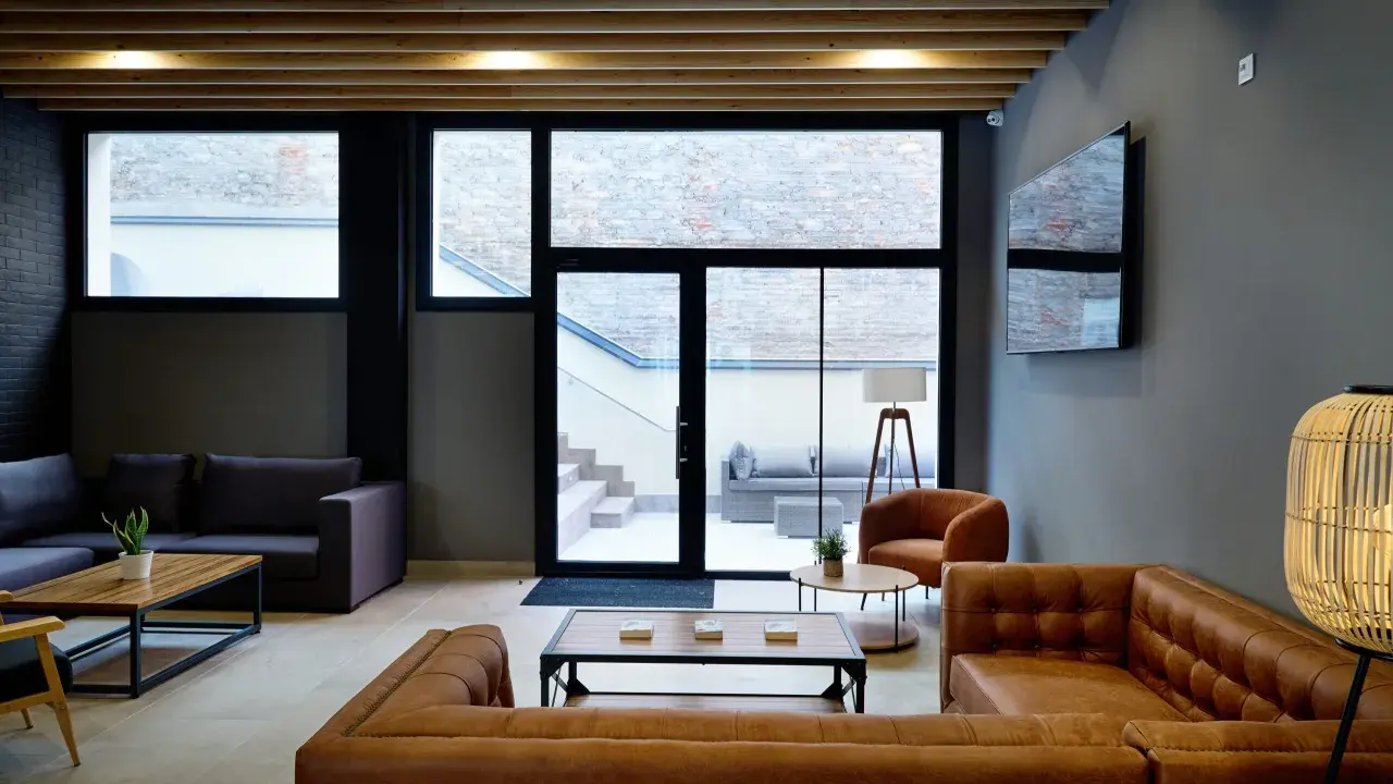 Sofá de cuero marrón y sillón naranja en la sala común de la residencia Xior Collblanc, con grandes ventanales que dan a un patio.