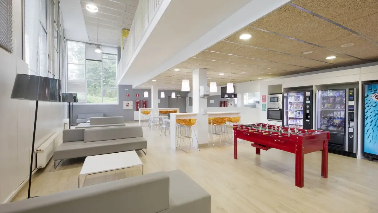 Sala común de una residencia estudiantil con sofás grises, futbolín rojo, máquinas expendedoras y mesas altas con taburetes naranjas.