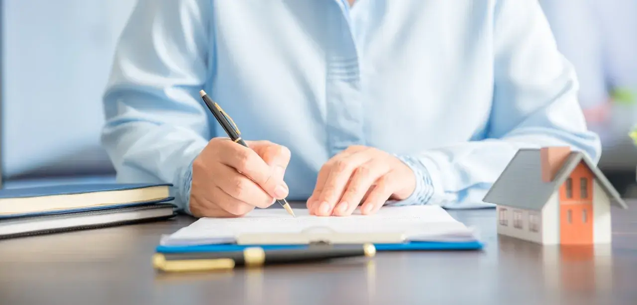 Persona firmando un documento en una mesa, donde se registra un contrato de alquiler. Hay una maqueta de casa y libros.