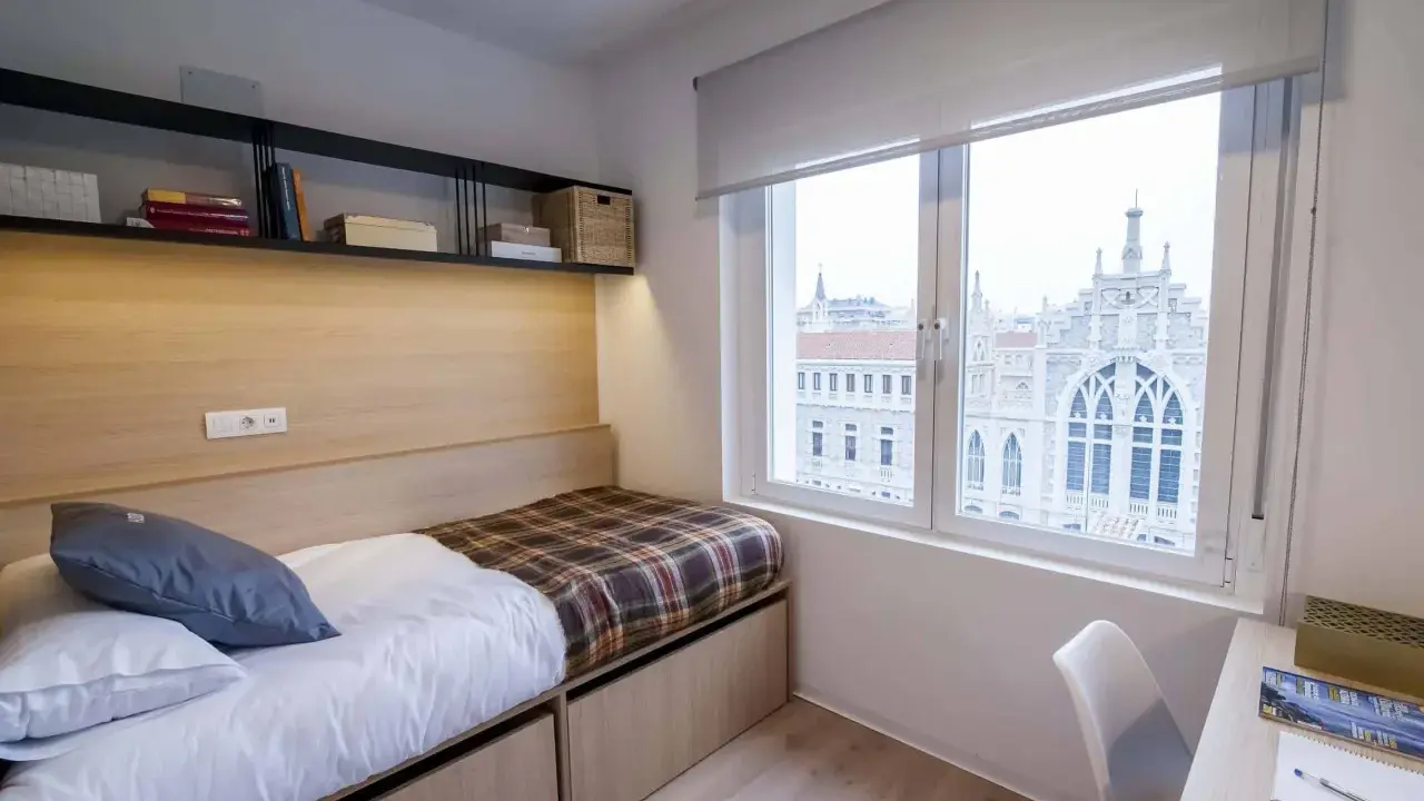 Habitación moderna con cama, estanterías y vistas a un edificio histórico en Xior Madrid Retiro.