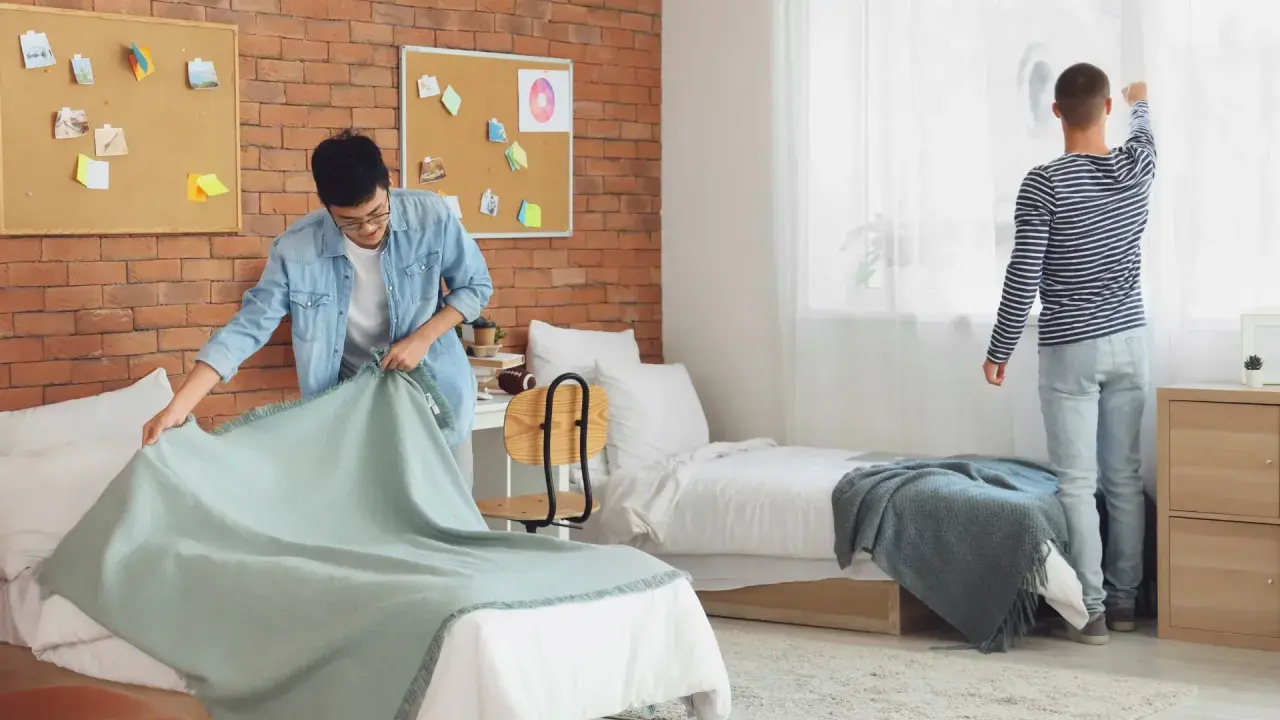 Dos jóvenes preparan su habitación de studea. Uno tiende una manta en la cama, el otro ajusta las cortinas.