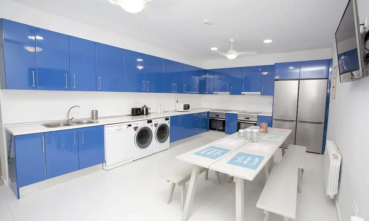 Cocina moderna con armarios azules brillantes, electrodomésticos y una mesa para compartir. Un espacio ideal para una **student factory** donde convivir y estudiar.