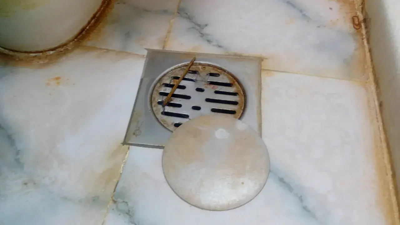 Desagüe de baño con rejilla metálica y tapa redonda. Se observa suciedad y sarro, indicando la necesidad de limpiar bote sifonico.