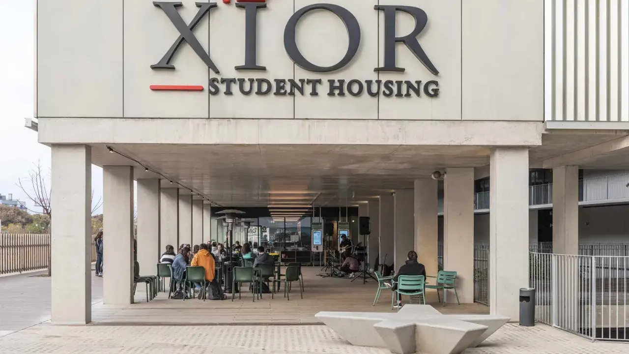 Estudiantes disfrutan en la **residencia Xior Picasso-Velázquez**. Un grupo toca música en vivo mientras otros charlan y comen al aire libre.