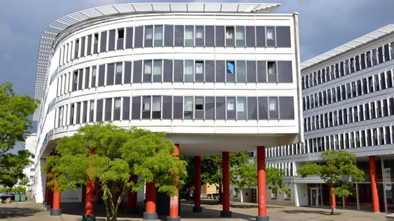 Edificio curvo blanco con ventanas oscuras y árboles verdes. Columnas rojas sostienen la estructura, creando un espacio crous.