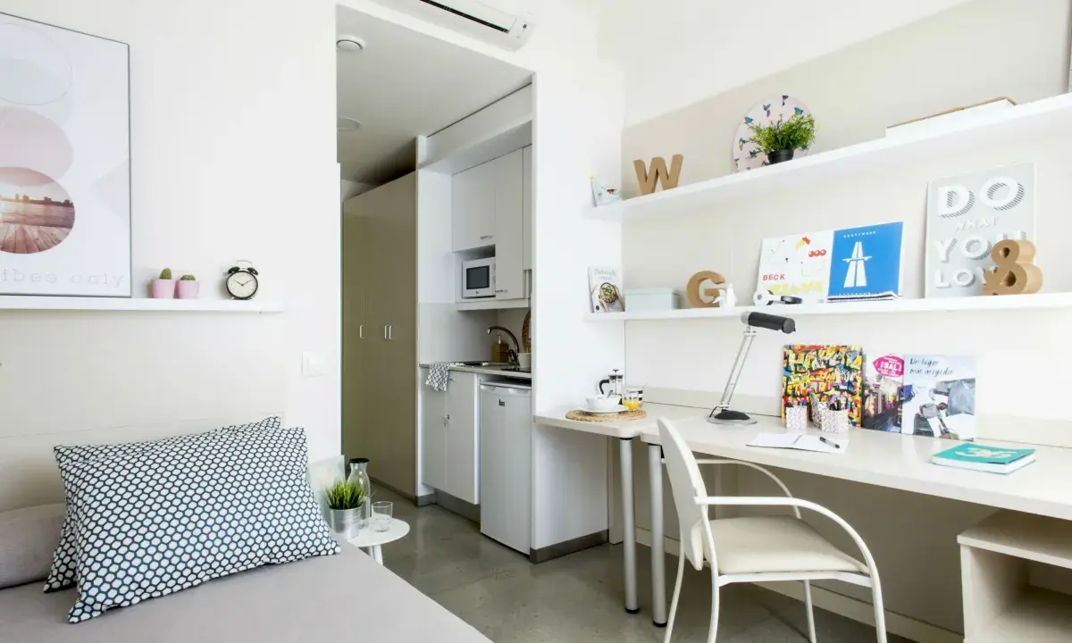 Estudio moderno en residencia La Salle: cama, escritorio con lámpara y estanterías, y pequeña cocina.