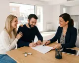 Pareja sonriente firma un contrato, quizás relacionado con el **articulo 2 LAU**, mientras una agente inmobiliaria les guía. Llaves sobre la mesa.
