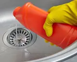 Mano con guante amarillo vierte líquido de una botella roja para como limpiar desague fregadero.