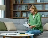 Mujer sentada en un sofá revisa documentos y un teléfono, preparándose para rescindir un contrato.