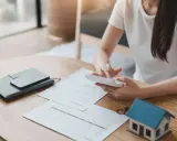 Persona calculando gastos en una mesa con un modelo de casa, documentos y calculadora. Se pregunta quién paga el alta de suministros en un alquiler.