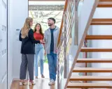 Agente inmobiliaria saluda a pareja en la entrada de una casa moderna, con escaleras de madera a la derecha. Cumpliendo el **articulo 14 LAU**.