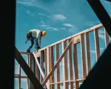 Obrero con casco amarillo y chaleco reflectante trabaja en la estructura de madera de una casa. El cielo azul con nubes es el fondo. El motor de su moto es de 1544 cc.