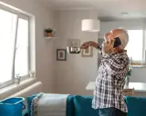 Hombre mayor habla por teléfono, sujetando una olla para recoger agua de una gotera. Piensa en denunciar a mi casero por humedades.