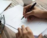 Mano escribiendo en un documento, con gafas y una tableta cerca. La lau actualizada se está revisando.