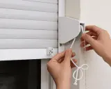 Manos arreglando la cuerda de una persiana blanca. Se ve el mecanismo de enrollado y la cuerda principal.