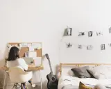 Joven trabajando en su escritorio en uno de los cuartos para estudiantes. Hay una guitarra, cama y fotos colgadas en la pared.