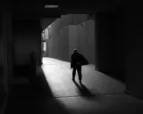 Hombre solitario camina por un pasillo oscuro, su figura se desvanece en la luz, un reflejo del precario significado de la existencia.