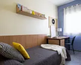 Habitación de estudiante en la carlos v residencia granada. Cama con cojines, escritorio con lámpara y estantería con libros.