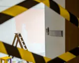 Escalera de madera y un taburete amarillo, listos para aplicar el **articulo 30 lau** en la pared. Cinta de precaución amarilla y negra.