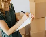 Mujer rubia con camiseta verde, revisando una lista en un portapapeles, rodeada de cajas de mudanza. El arte 19 lau inspira la organización.