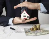 Manos protegen una casa modelo sobre un contrato de alquiler con avalista, junto a dinero y gráficos.