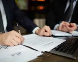Dos hombres de negocios revisan un contrato. Uno firma el documento, mientras el otro observa. Podría tratarse de un acuerdo legal, o quizás de un proceso de embargo por impago de alquiler.
