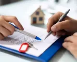 Manos firmando un documento, con llaves y una maqueta de casa. Podría ser un desistimiento de contrato de alquiler.