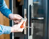 Man con camisa de mezclilla instala un cilindro de cerradura en una puerta, resolviendo un problema de comodato precario.