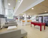 Sala común de una residencia estudiantil con sofás grises, futbolín rojo, máquinas expendedoras y mesas altas con taburetes naranjas.