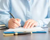 Persona firmando un documento en una mesa, donde se registra un contrato de alquiler. Hay una maqueta de casa y libros.