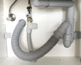 El fregadero pierde agua por abajo. Tuberías grises y mangueras flexibles bajo el lavabo, con una fuga visible.