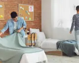Dos jóvenes preparan su habitación de studea. Uno tiende una manta en la cama, el otro ajusta las cortinas.