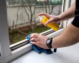 Persona rocía un limpiador en un marco de ventana y lo limpia con un paño azul. Es un paso para quitar moho paredes por condensación.