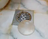 Desagüe de baño con rejilla metálica y tapa redonda. Se observa suciedad y sarro, indicando la necesidad de limpiar bote sifonico.