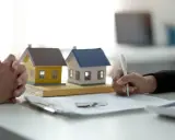 Manos entrelazadas junto a maquetas de casas y un bolígrafo firmando un contrato privado de alquiler.
