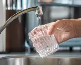 Mano sujetando un vaso de cristal bajo un grifo, llenándolo de agua. Un momento especial para dar de alta el agua por primera vez en un nuevo hogar.