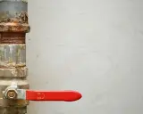 Llave de paso agua antigua, oxidada y con palanca roja, controlando el flujo.