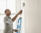 Hombre pintando pared blanca, resolviendo quién paga la pintura en un piso de alquiler. Escalera azul al fondo.