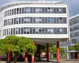 Edificio curvo blanco con ventanas oscuras y árboles verdes. Columnas rojas sostienen la estructura, creando un espacio crous.