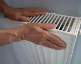 Manos sobre un radiador blanco, sintiendo el calor. Quizás se pregunte porque suenan los radiadores.