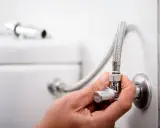 Mano ajustando la llave de paso del agua, que es comunitaria, conectada a una manguera flexible y al inodoro.