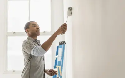 Hombre pintando pared blanca, resolviendo quién paga la pintura en un piso de alquiler. Escalera azul al fondo.